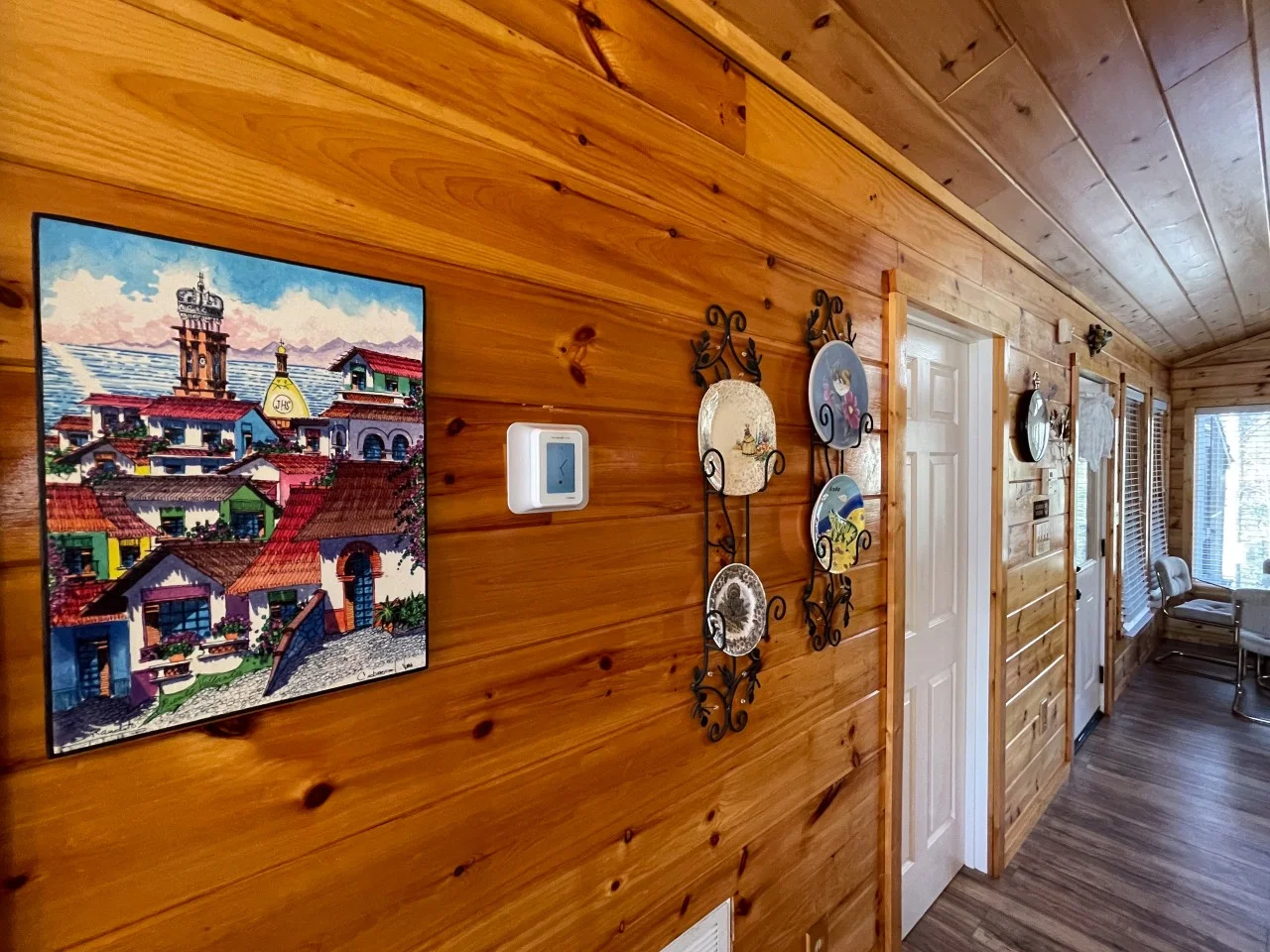 Cabin hallway with honey-colored wood paneling and colorful plates, folk art on a wall-mounted TV, a cabin hot tub in winter
