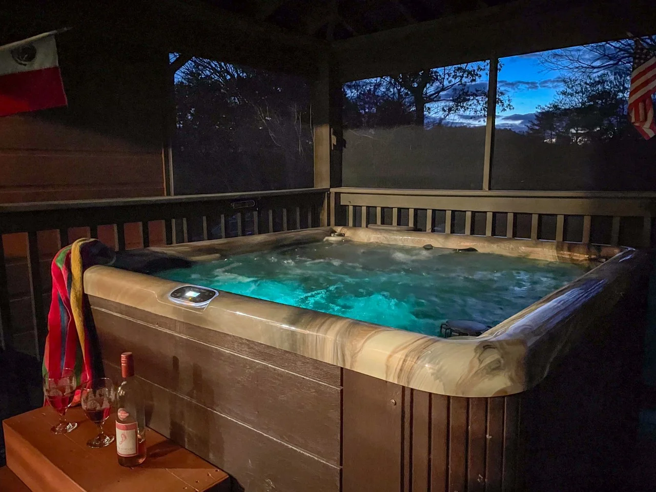 Illuminated turquoise hot tub on a covered deck at dusk with Smoky Mountain view through large windows at a Pigeon Forge, TN